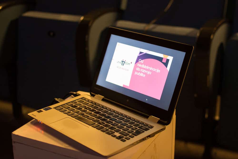 Laptop je na drvenoj površini. Na levoj strani ekrana laptopa nalazi se logo Uhvati film, dok na desnoj strani piše natpis: Od nediskriminacije do razvoja publike. U pozadini su obrisi bioskopskih stolica.