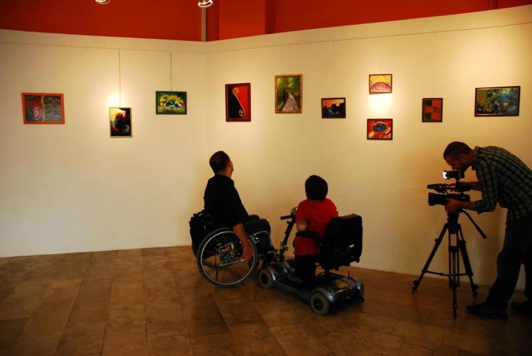 Na zidovima su slike a ispred dve osobe ( korisnici invalidskih kolica) posmatraju