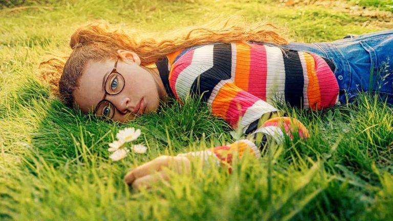 Devojka leži na travi, licem okrenuta ka zemlji. Zamišljena je i drži cvet u rukama.