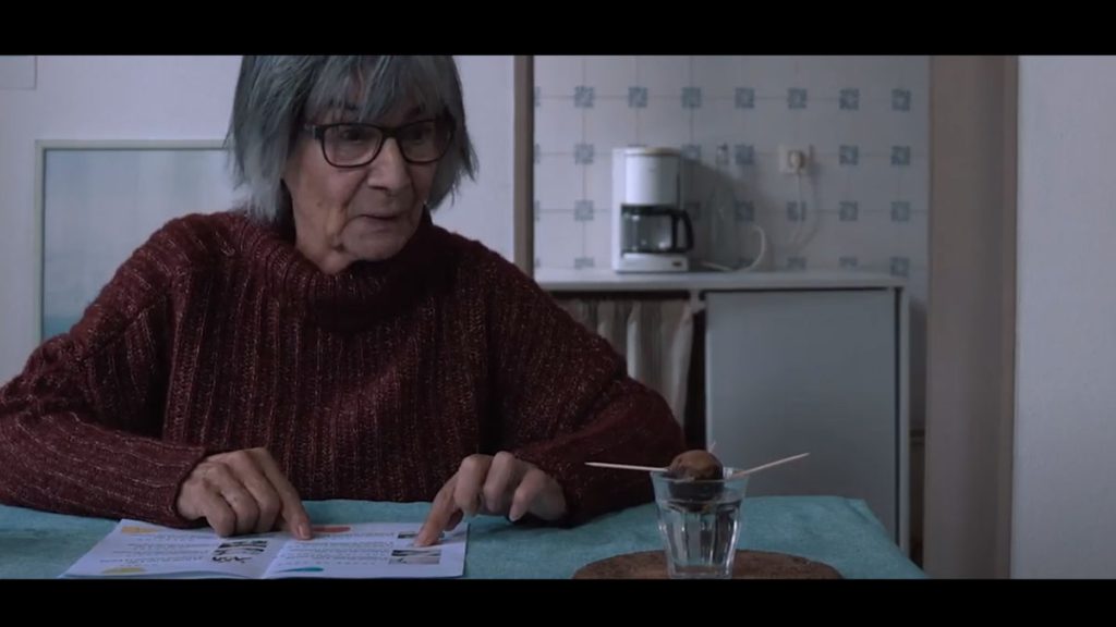 Starija žena sedi za stolom. Na stolu je i otvorena brošura i u čaši kugla nečega sa zabodenim čačkalicama. Žena gleda u čašu i izgled kao da se obraća čaši.