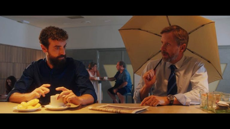 Dvojica muškaraca za stolom u kafiću. Ispred njih su kafa, čaše i novine. Jedan od njih nosi kišobran. Iza njih vide se drugi ljudi u kafiću.