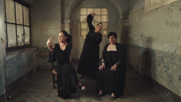 Tri žene u prostorijama kamenog zamka. Dve sede, a žena između njih stoji sa podignutom jednom rukom.